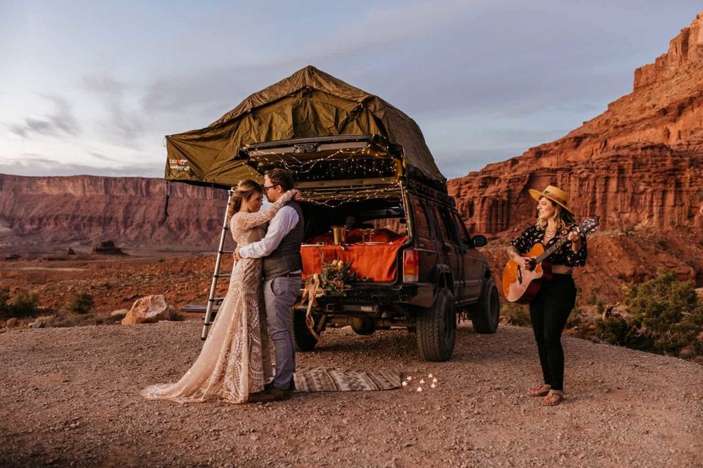 A couple shares a slow dance by the back of their jeep as a vendor plays guitar and sings for them.