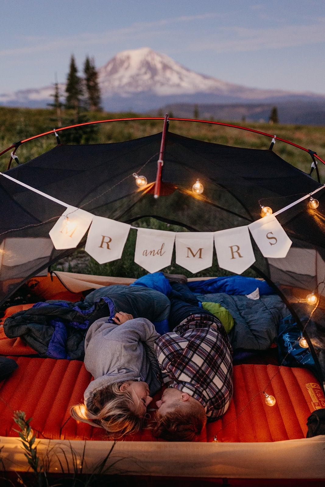 A couple lays in a tent together near a mountain with their just married sign.