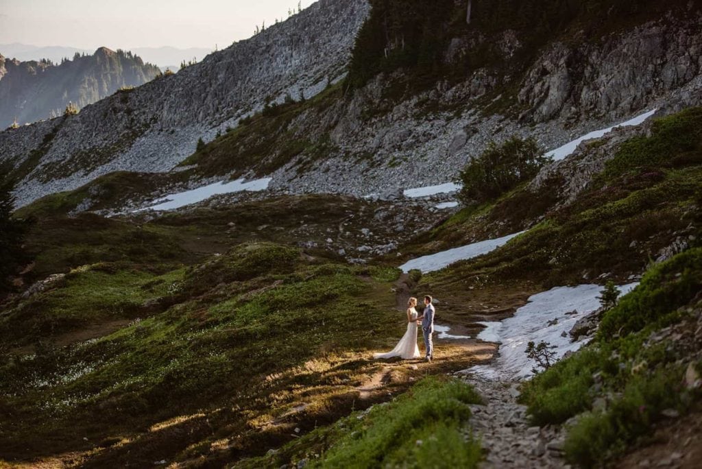 A couple in the sunlight standing in the mountains near their camp in their wedding attire.