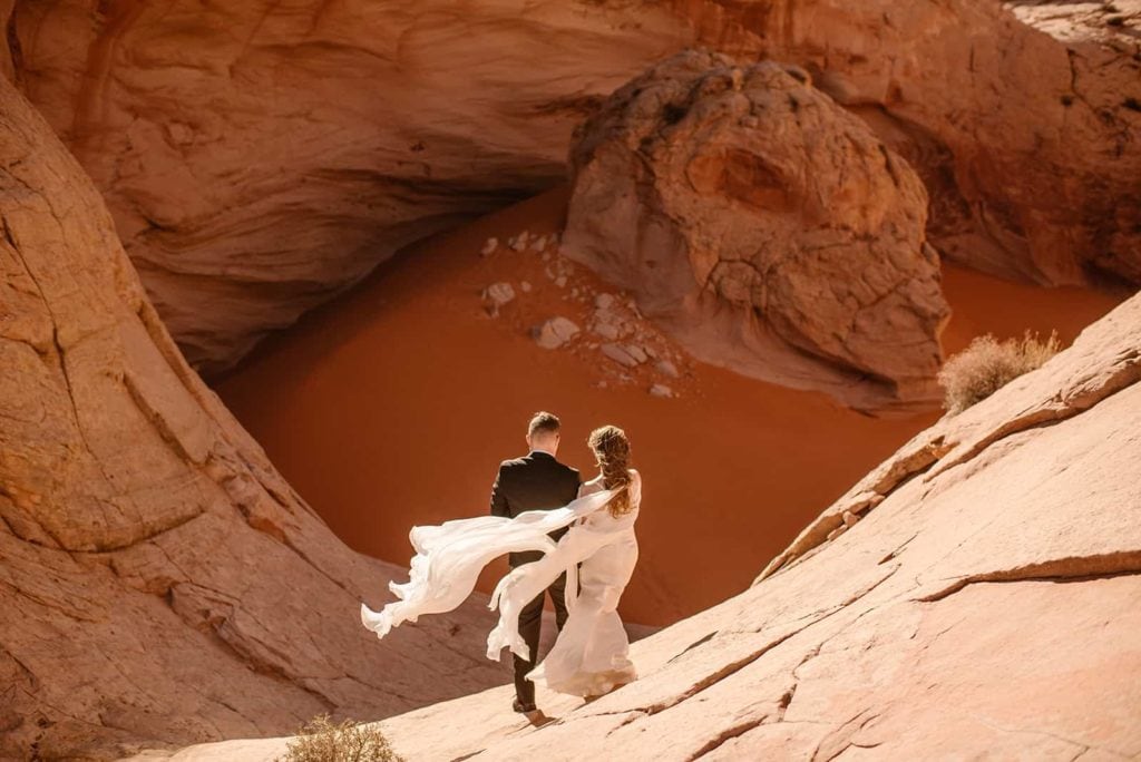 A couple faces a canyon filled with sand as the wind catches the brides dress. 