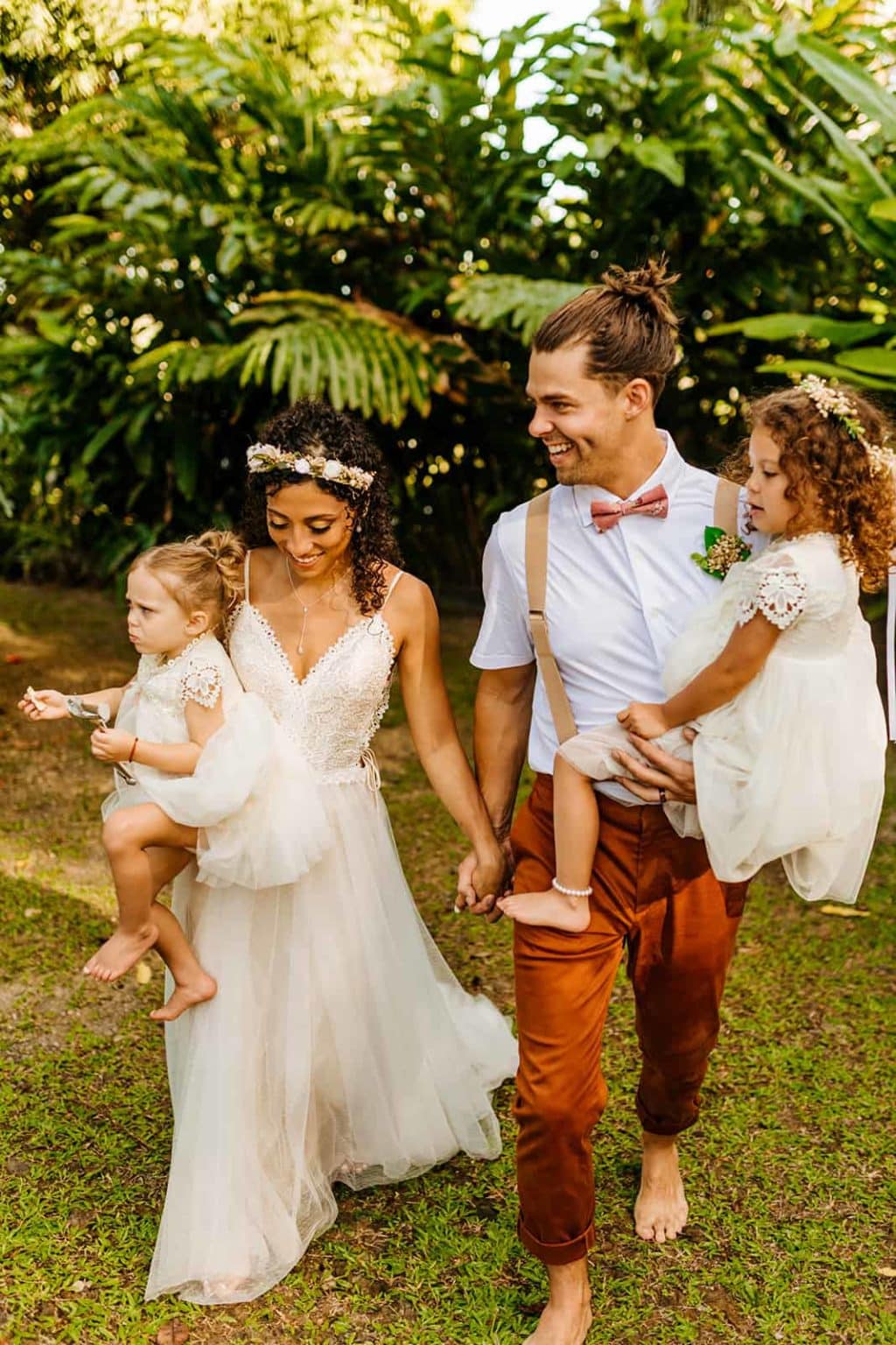 A bride and groom walk through a field with their kids.
