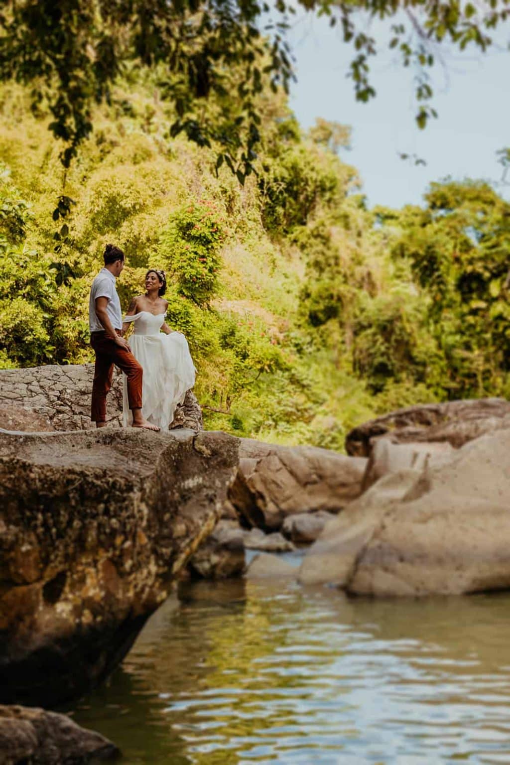 A bride and groom count down together when to jump off of a rock into the water.