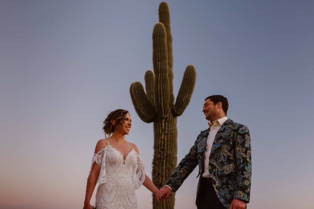 A bride and groom hold hands staring at each other with a large cacti behind each other.