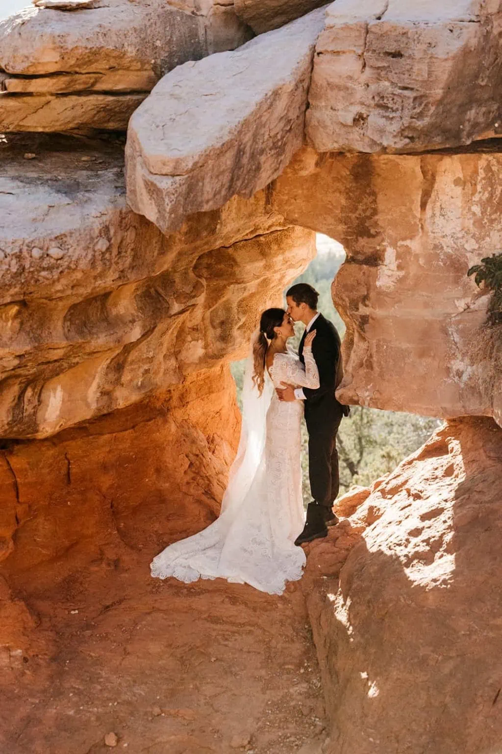 A bride and groom stand together in a cave and the groom kisses the brides forehead.
