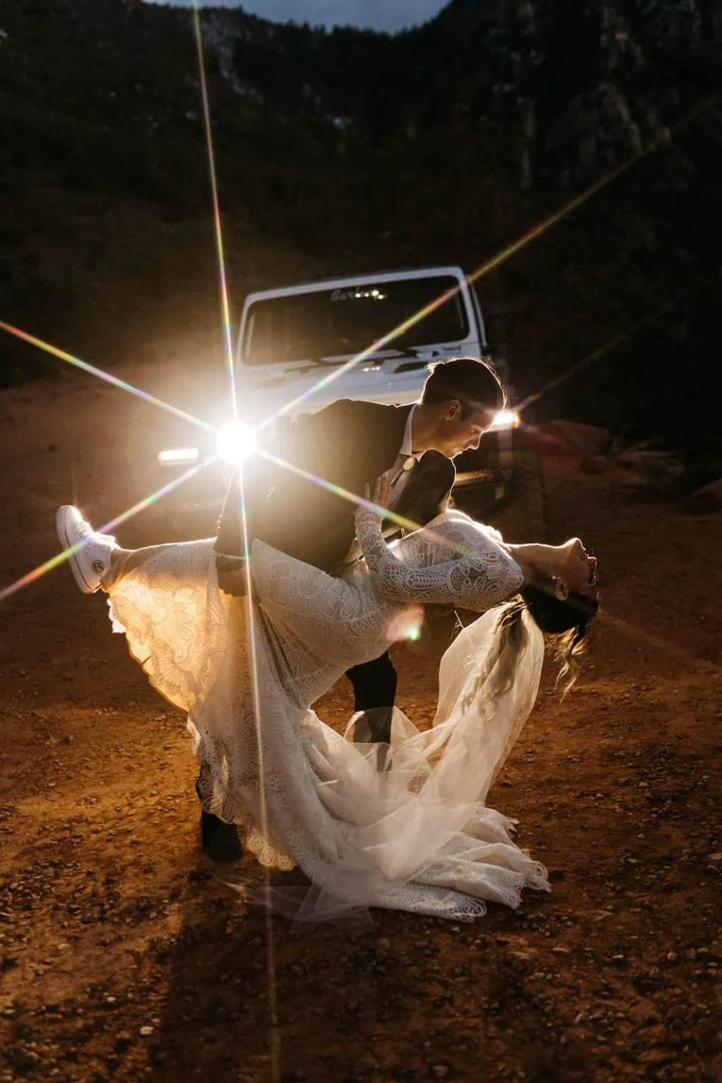 A groom dips his bride back in front of the lights of a jeep.