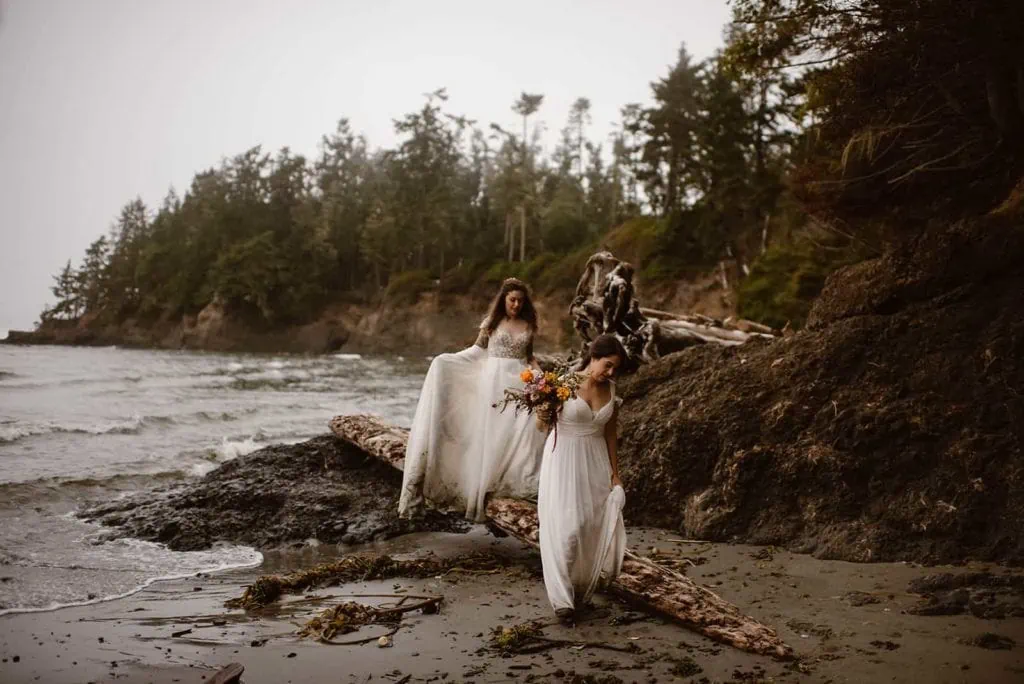 Two brides walk along a long by the Olympic coast on a cloudy day.