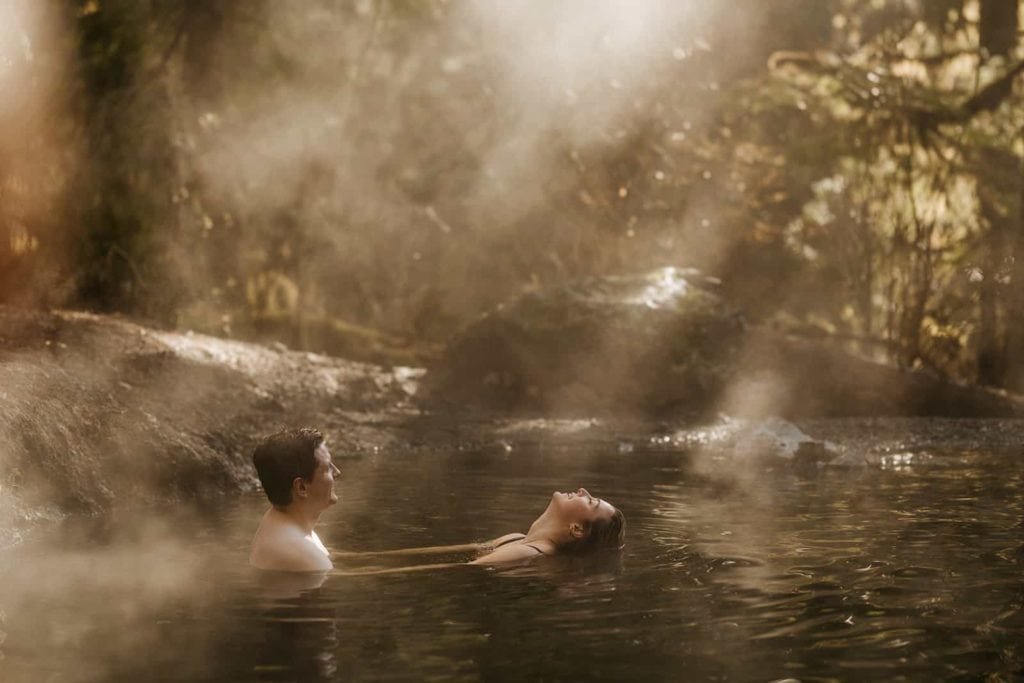 A girl leans her head back into the hot spring as the sun creates a mist around the couple as they are surrounded by forest. 