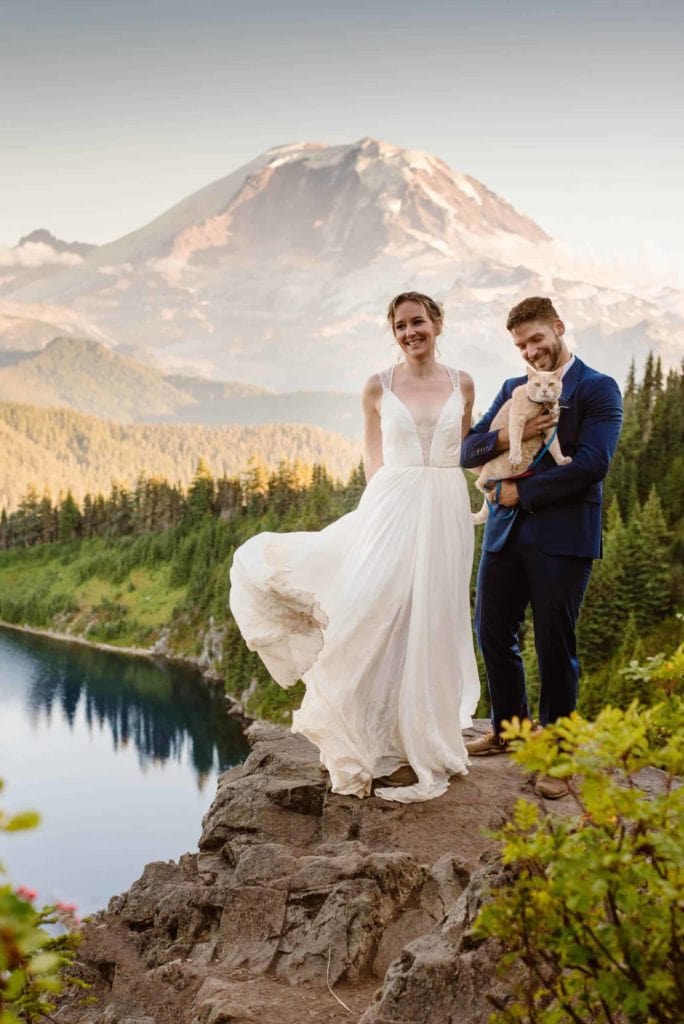A bride and groom stand together while holding their cat with a view of Mount Rainier behind them.