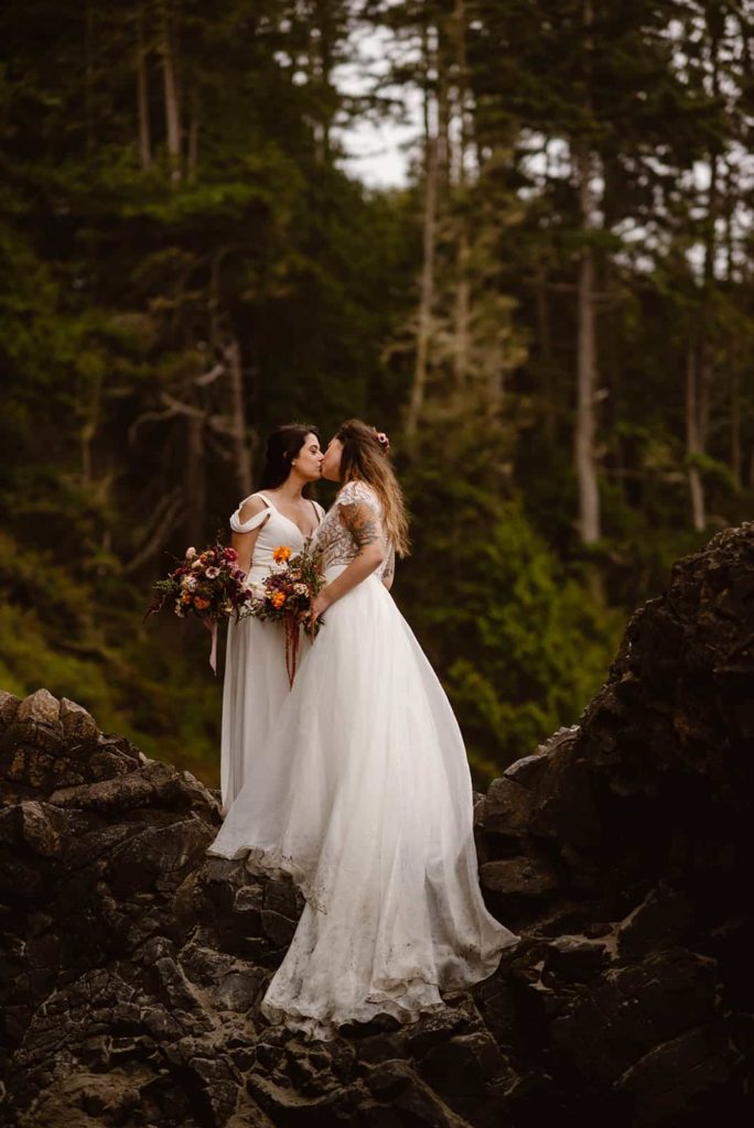 two brides kissing each other in the woods, both wearing wedding dresses.