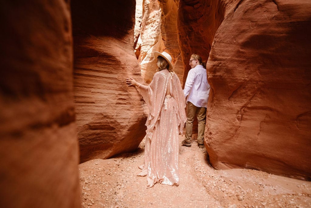 A man and woman walk through a slot canyon in Moab as they feel the texture of the rocks. 