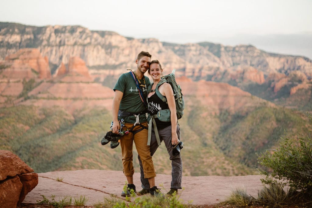 A couple stands together holding their climbing gear and one of them wearing a backpack up high on a ledge with red rock views behind them.