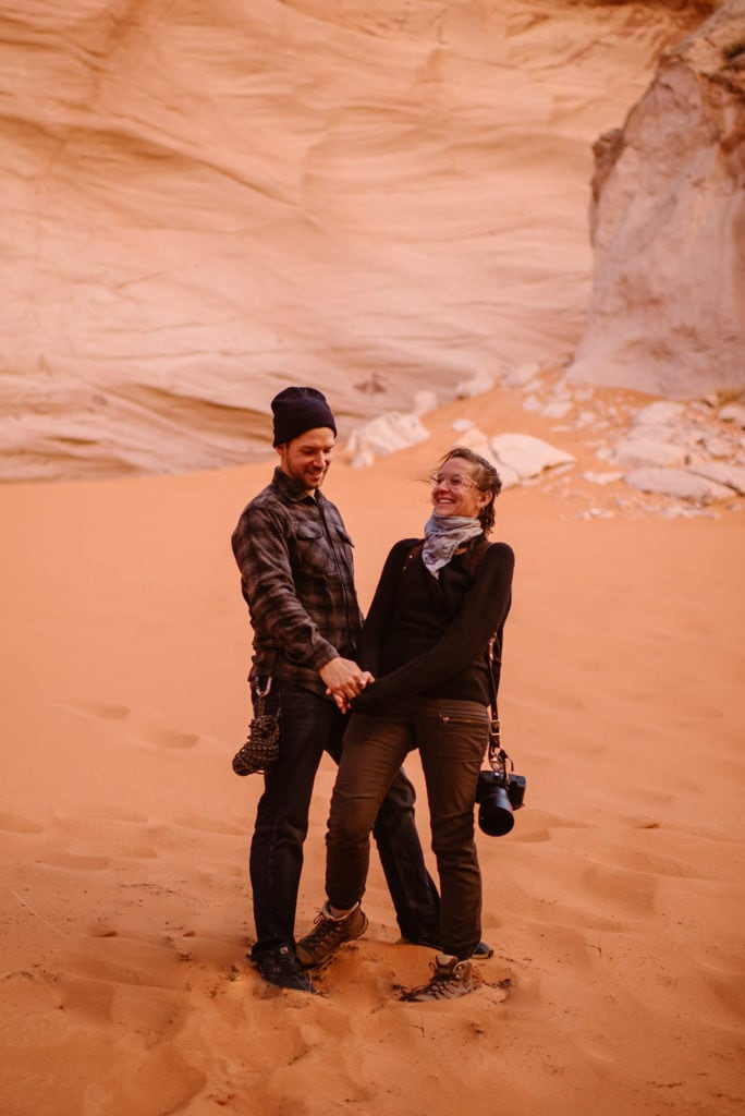 A woman and a man hold hands smiling towards each other standing in sand.