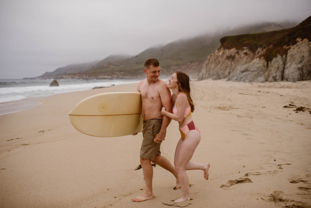 a bride and groom run along the beach, smiling widely. he wears beige board shorts, while she wears a pink and red bikini. he's carrying a surfboard.