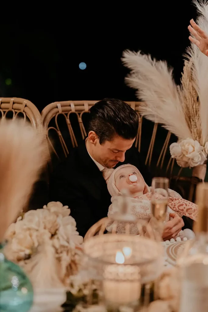 A groom holding a baby at the dinner table.