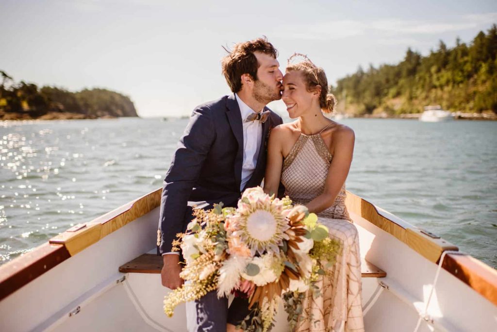 a bride and groom are in a boat on a sunny day. the groom is kissing his bride's head. she is holding her bouquet.