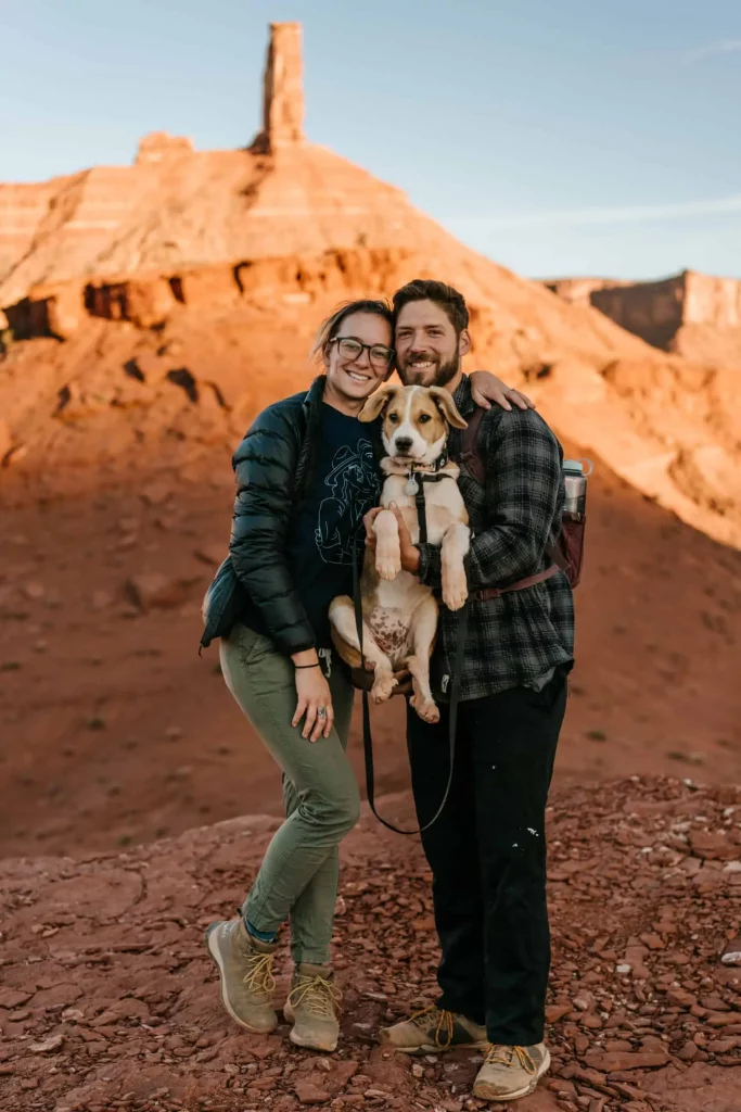 A man and woman stand together holding a puppy during sunrise in Moab Utah