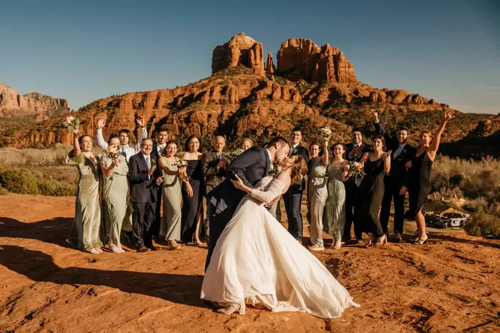 A groom dips his bride back for a kiss while their guests celebrate in the background.