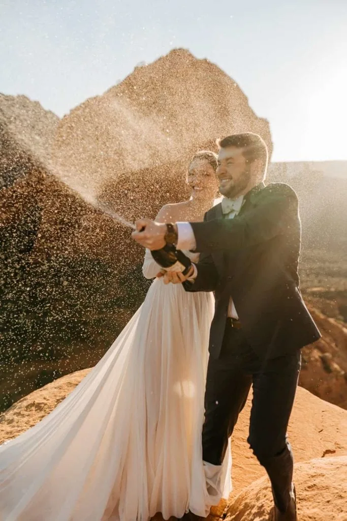 A groom sprays a bottle of champagne at sunrise in Sedona with his bride by his side.