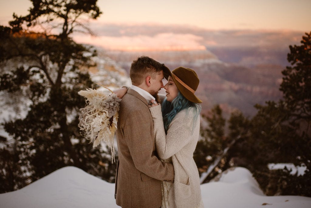 a couple embrace, touching noses at the Grand Canyon with snow surrounding them.