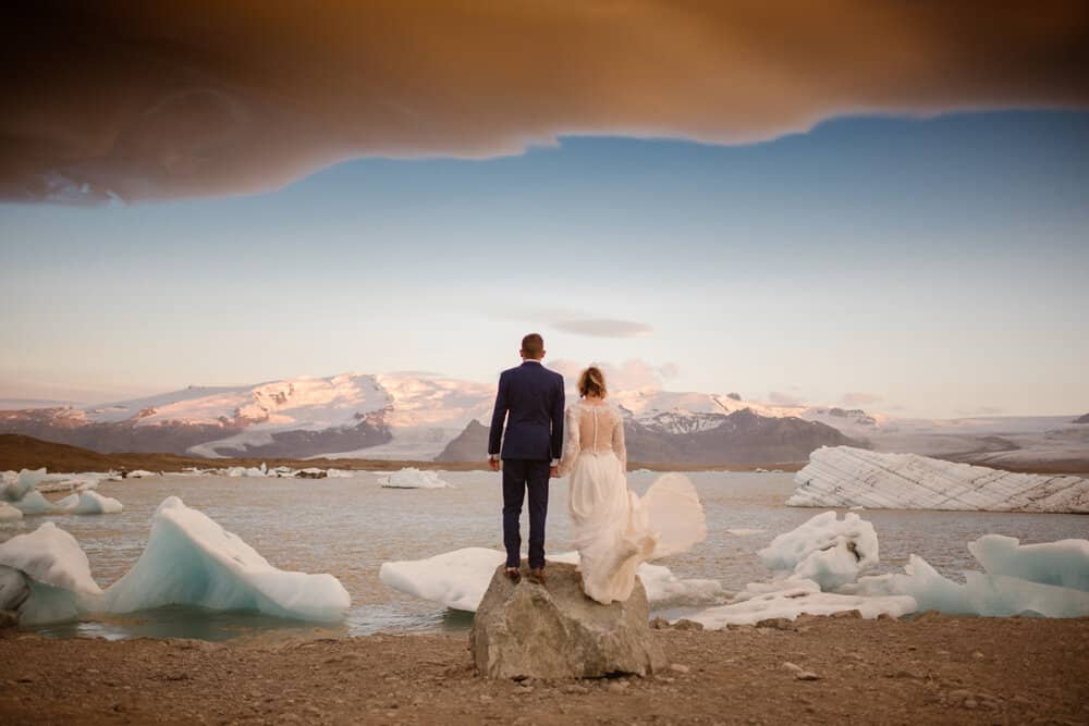 bride and groom overlook iceland's glaciers and water of Iceland hand in hand. her dress is white and blows in the wind, while his suit is black.