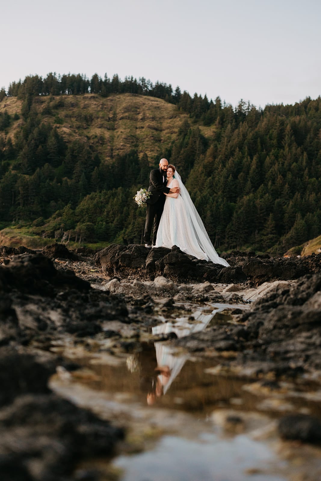 A couple stands together where they got engaged while on their wedding day with their reflection in the water. 