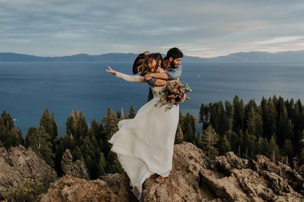 a couple strike a pose on the top of lake Tahoe's mountains. the ocean and mountains are in the background, below them.