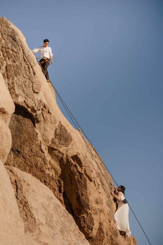 A couple rock climbs together in their wedding attire on a sunny day in Joshua Tree. 