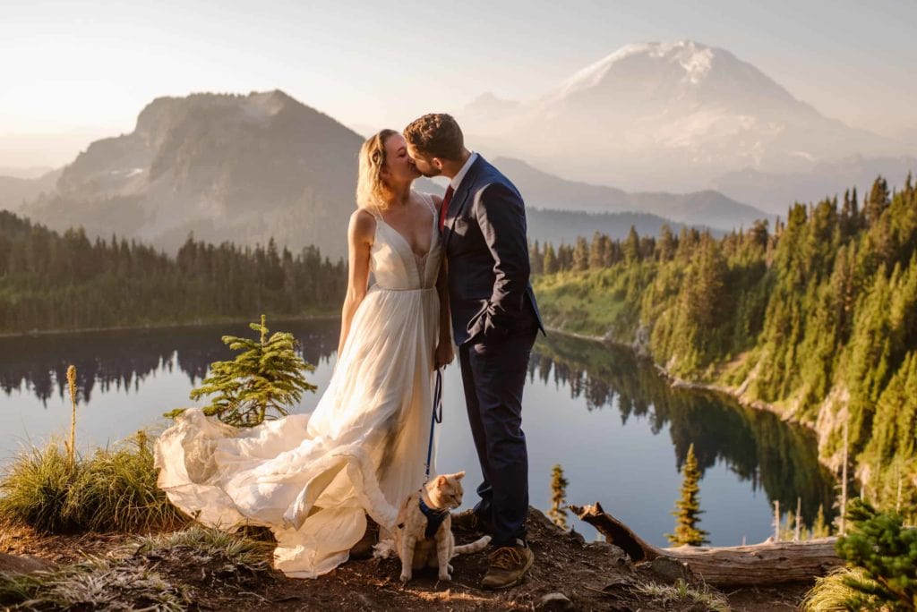 A couple kiss in the morning light as their cat stands behind them with epic mountains views all around.