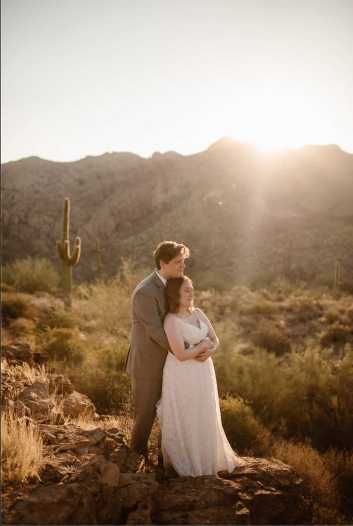 a groom holds his wife from behind as they look off into the distance. there is a cactus in the background.