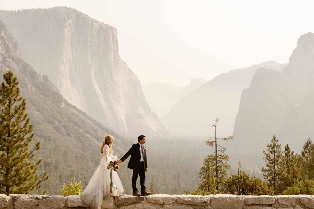 A couple walks along a rock wall in the Yosemite Valley on a hazy day showcasing all of the peaks in the park. 