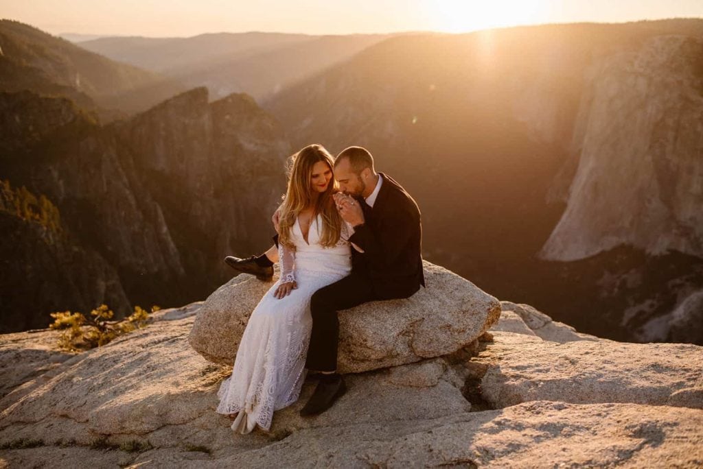 a couple sitting on a rock overlooking Yosemite. the man is kissing his bride's hand.