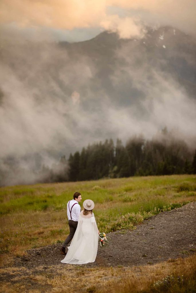 a couple walking along the hillside, fog blowing along the mountains. She is wearing a dress and sun hat, while he wears a white shirt and suspenders.