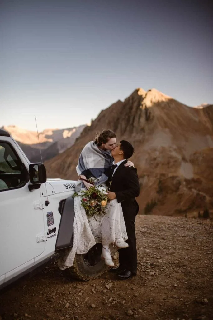 A bride and groom share a kiss while in the mountains and the bride sits on her jeep.