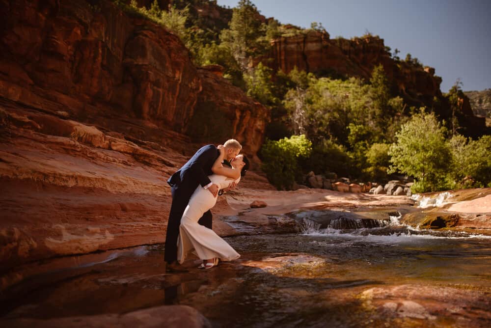 A groom gives his bride a dip kiss as they stand in a low river. 