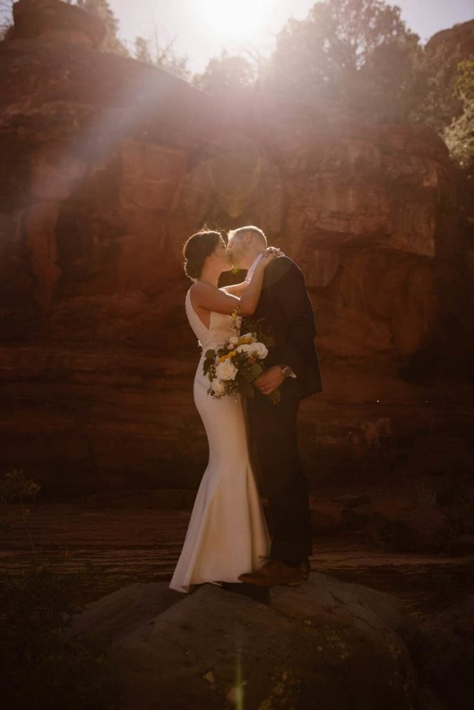 A couple shares a kiss as they stand on a rock with red rock walls behind them. 