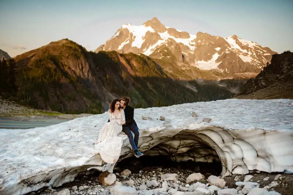 A bride and groom sit on a glacier together with mountains behind them.