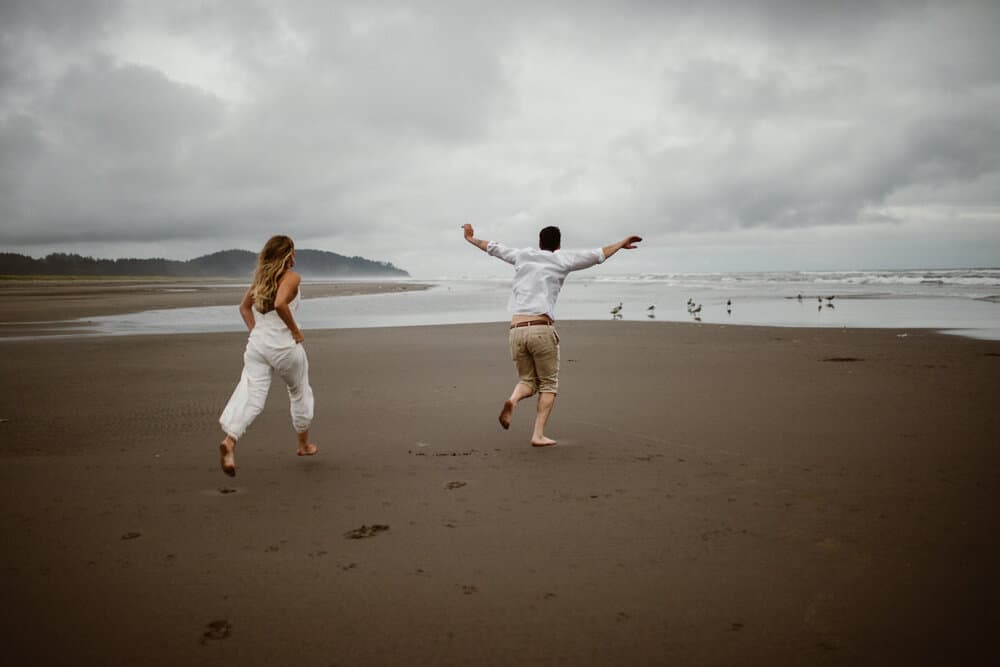 A bride and groom run together towards the ocean.