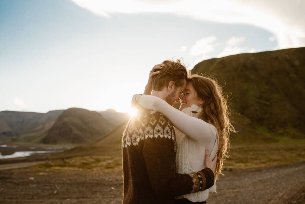 A woman embraces her partner as the sun peaks from around them and the mountains. 
