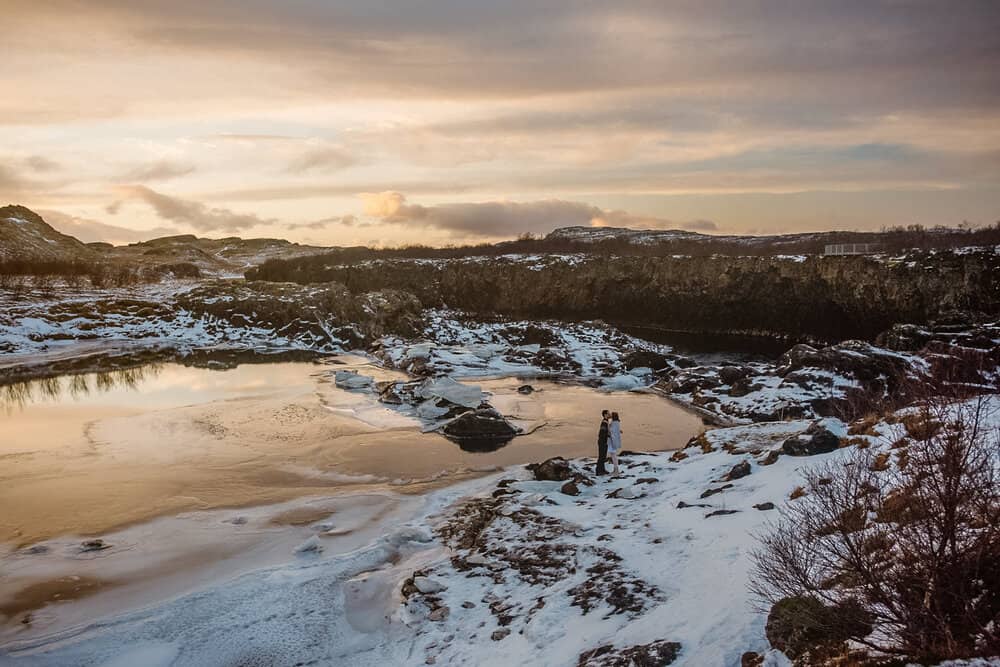 A couple stands by a body of water surrounded by snow on a Winter day in Iceland on the morning of their elopement. 