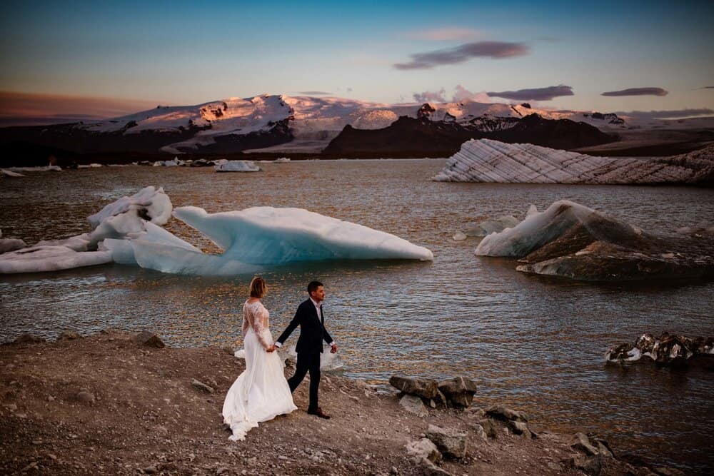 A groom leads his bride down the shore line of a lake in Iceland with iceburgs floating in it at midnight. 