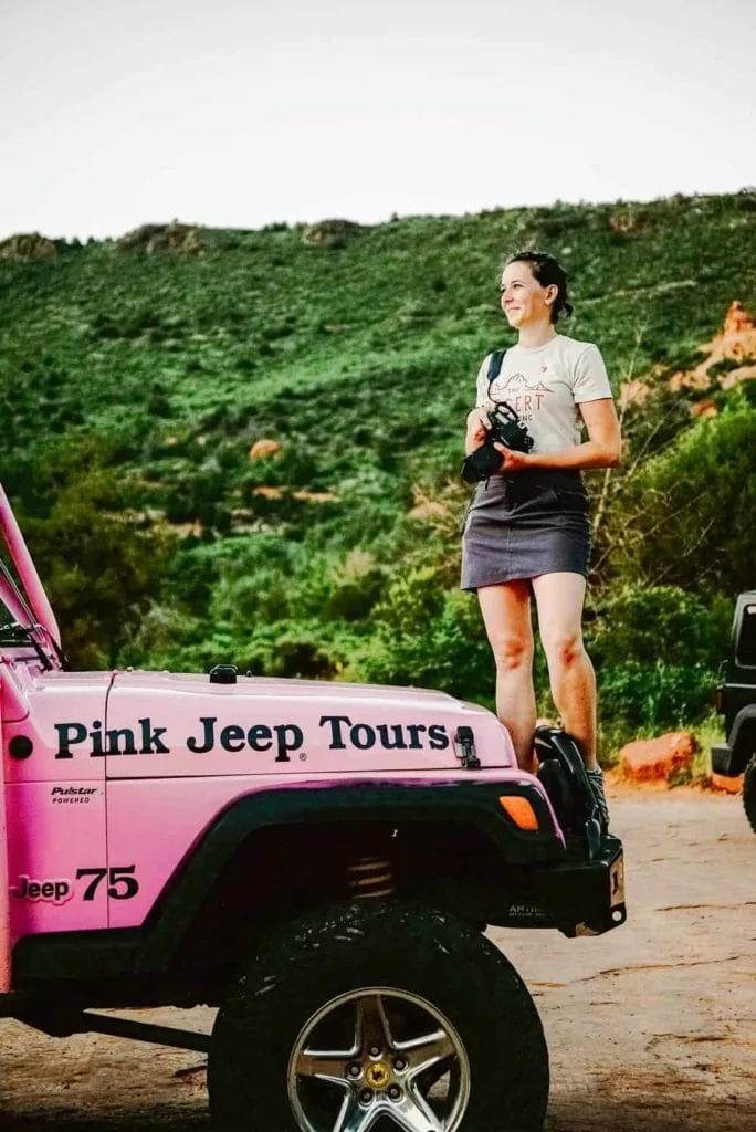 A girl stands on a pink jeep with a camera in her hand.