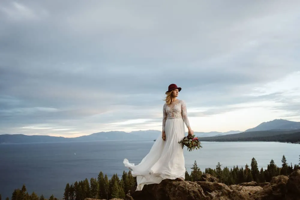 A brides stands overlooking lake tahoe on a windy day in a red hat.