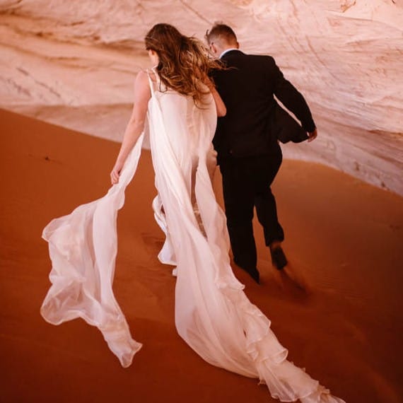 a bride and groom walking through a canyon, sand red as they walk. the bride's dress trails behind her.