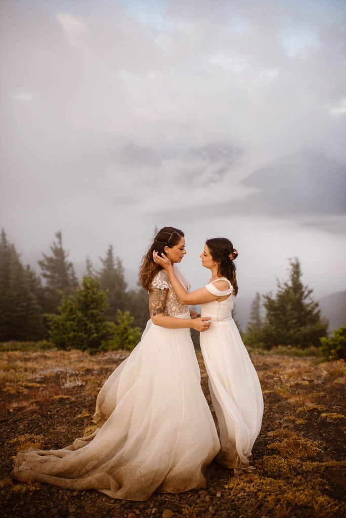 Two brides longingly share a moment together on top of a mountain in the clouds. 