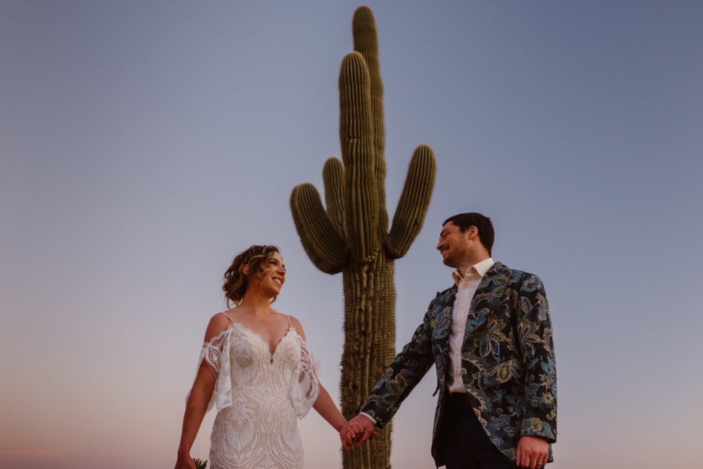A couple holds hands and admires each other as a tall cacti stands behind them. 