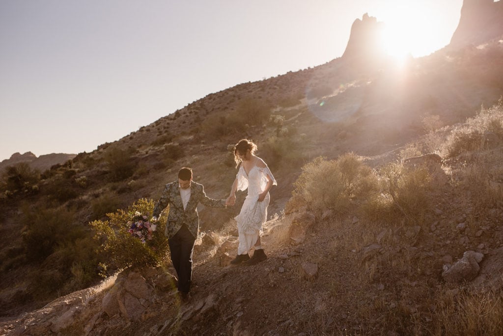 A groom holds his brides hand while they carefully walk down the rocky trail with the sun coming up behind them. 