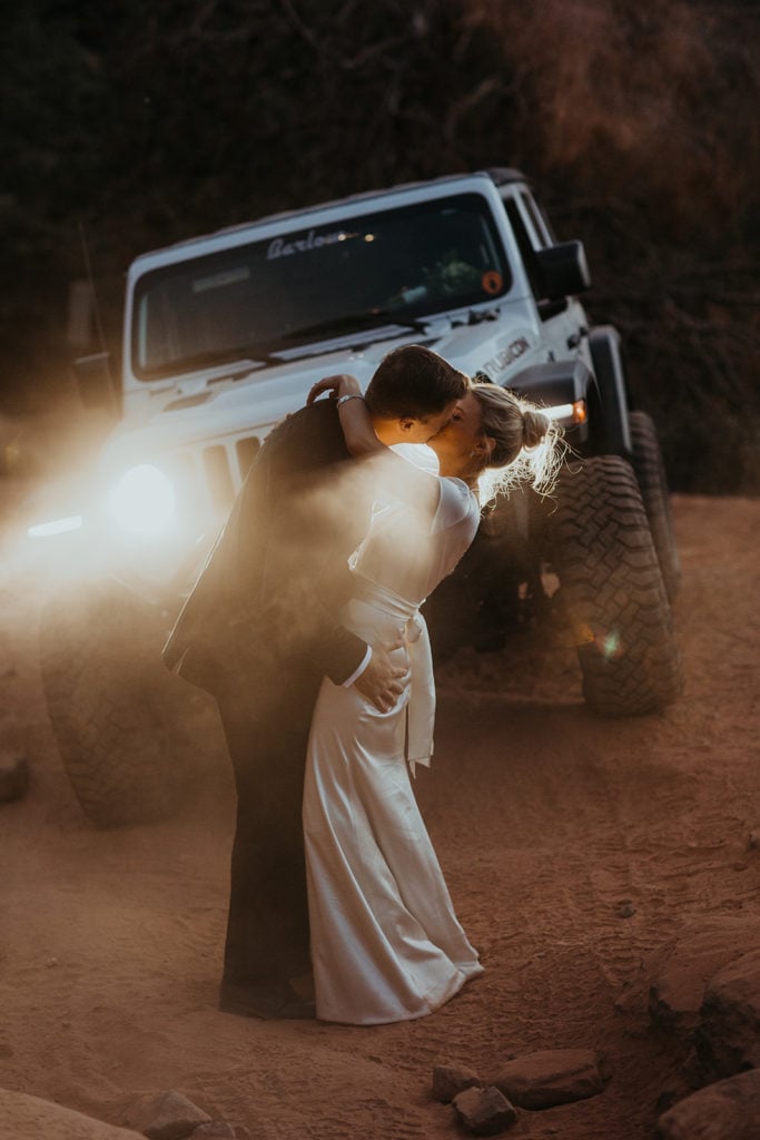A man and woman kiss in the dust and light of their jeep headlights before starting off roading back down the mountain.