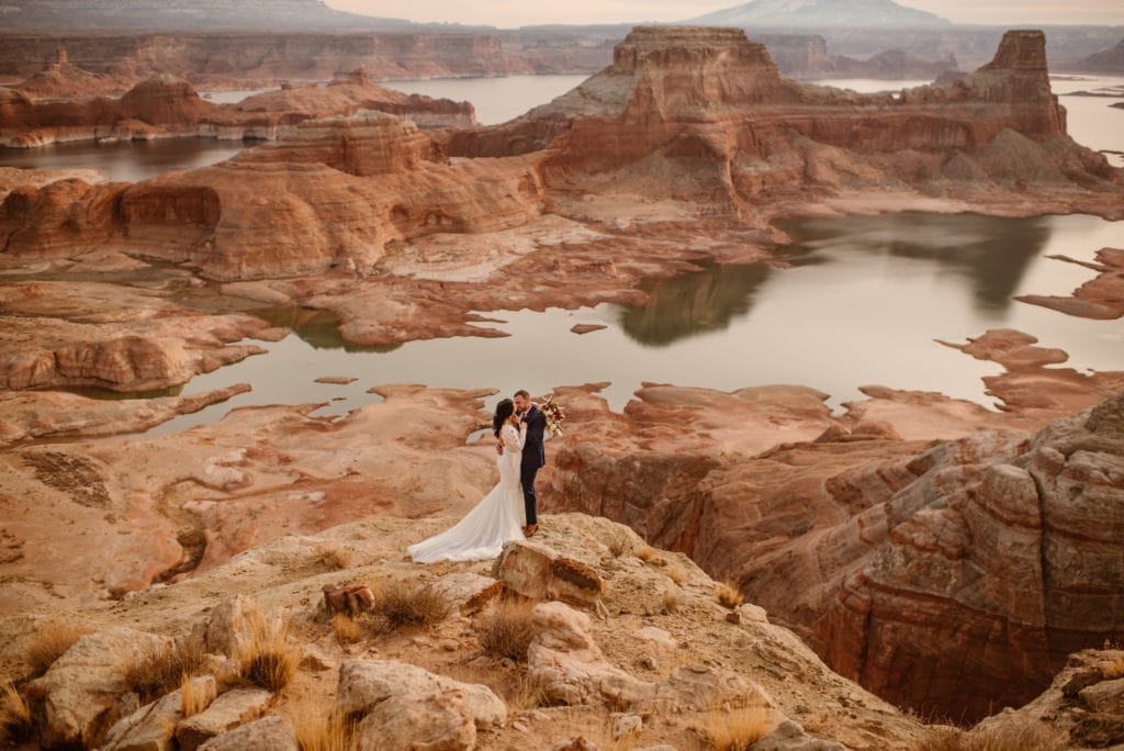 A couple holds each other close on a winter morning overlooking red rock canyons and a lake. 