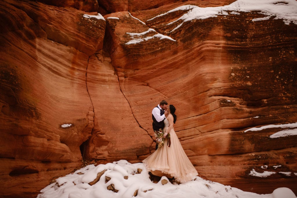 a couple kissing at the Grand Canyon. she wears a pink gown and carries a bouquet, while he wears a black vest and white shirt, embracing her face.