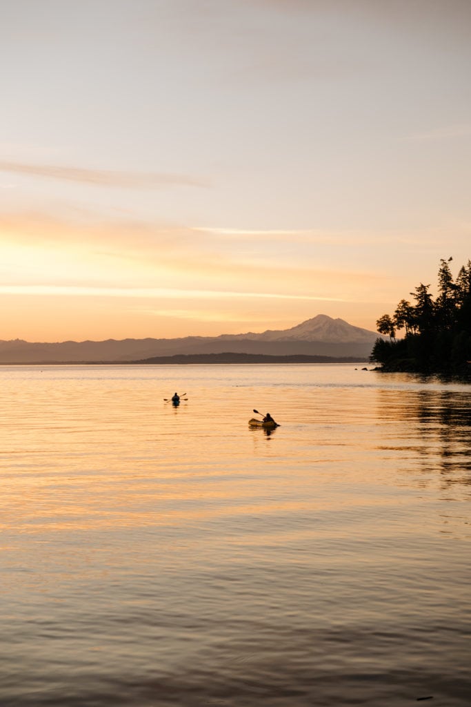 Two people on kayaks in the bay at sunrise with a mountain view behind them.